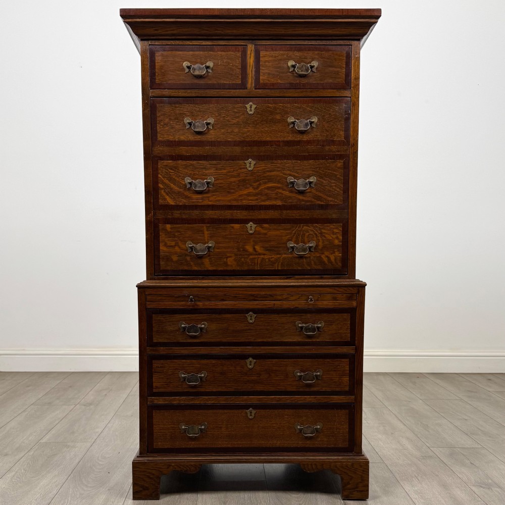 A tall early 20th-century oak chest on chest featuring multiple drawers with crossbanded borders and brass drop handles set on bracket feet.