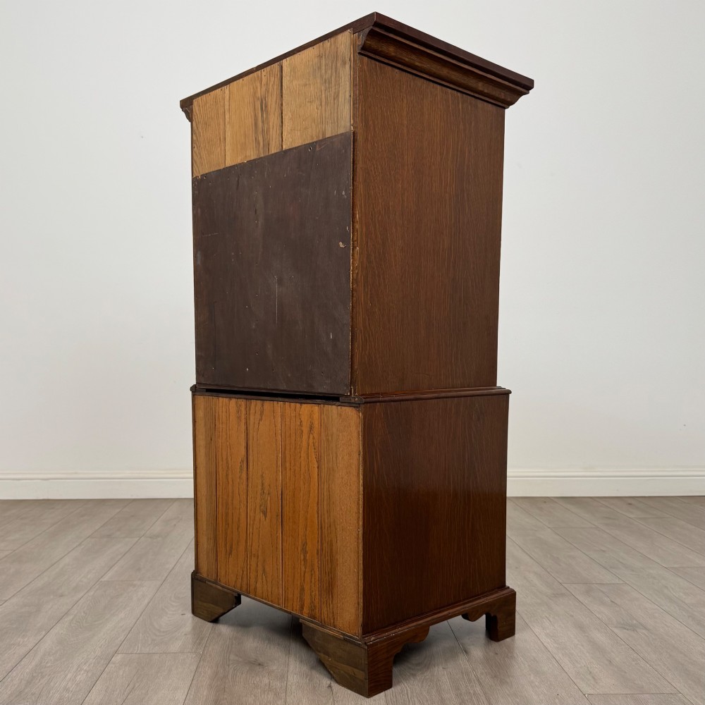 A tall early 20th-century oak chest on chest featuring multiple drawers with crossbanded borders and brass drop handles set on bracket feet. View VII