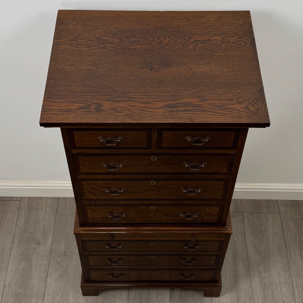 A tall early 20th-century oak chest on chest featuring multiple drawers with crossbanded borders and brass drop handles set on bracket feet. View X