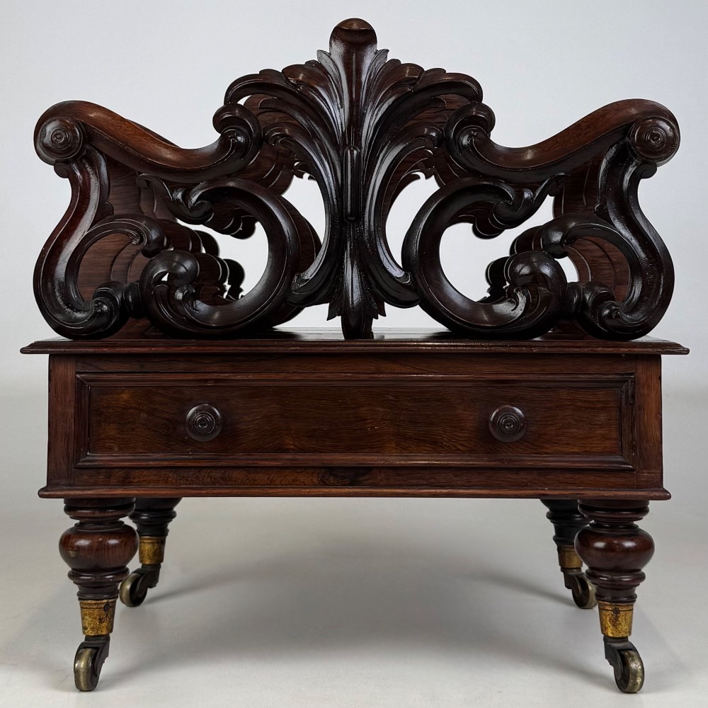 A dark rosewood Victorian canterbury with central acanthus carving, double scrollwork dividers, a single lower drawer, turned legs, and brass castors.