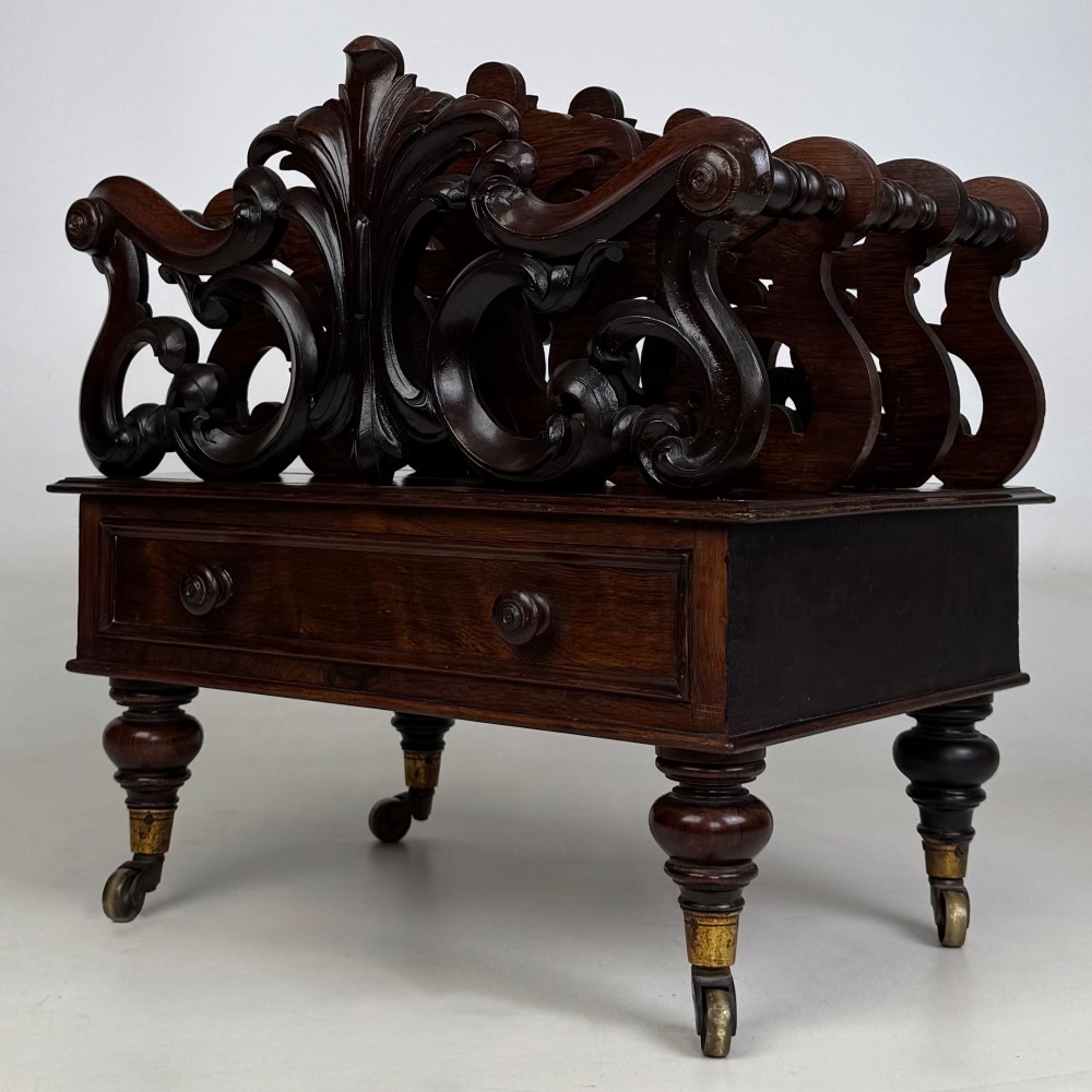 A dark rosewood Victorian canterbury with central acanthus carving, double scrollwork dividers, a single lower drawer, turned legs, and brass castors. View II