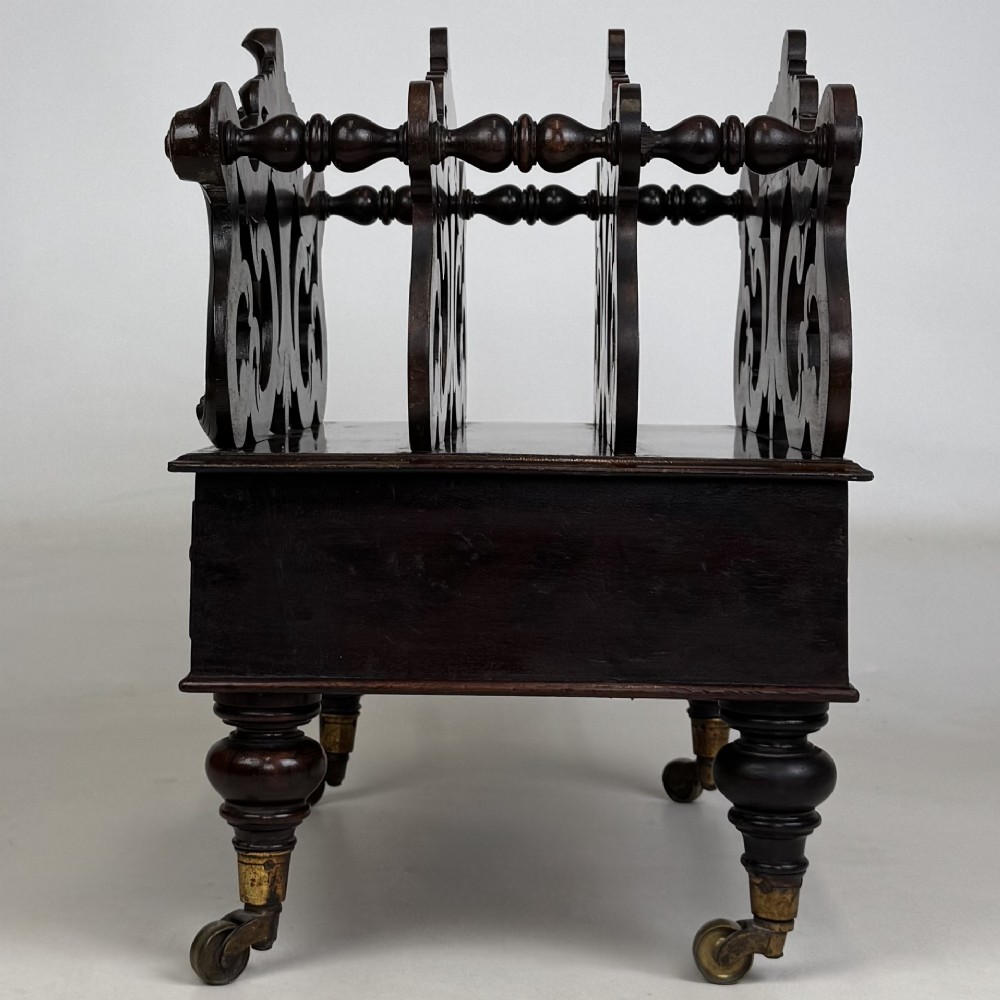 A dark rosewood Victorian canterbury with central acanthus carving, double scrollwork dividers, a single lower drawer, turned legs, and brass castors. View III