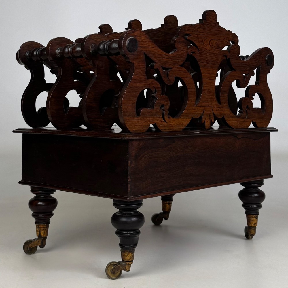 A dark rosewood Victorian canterbury with central acanthus carving, double scrollwork dividers, a single lower drawer, turned legs, and brass castors. View IV