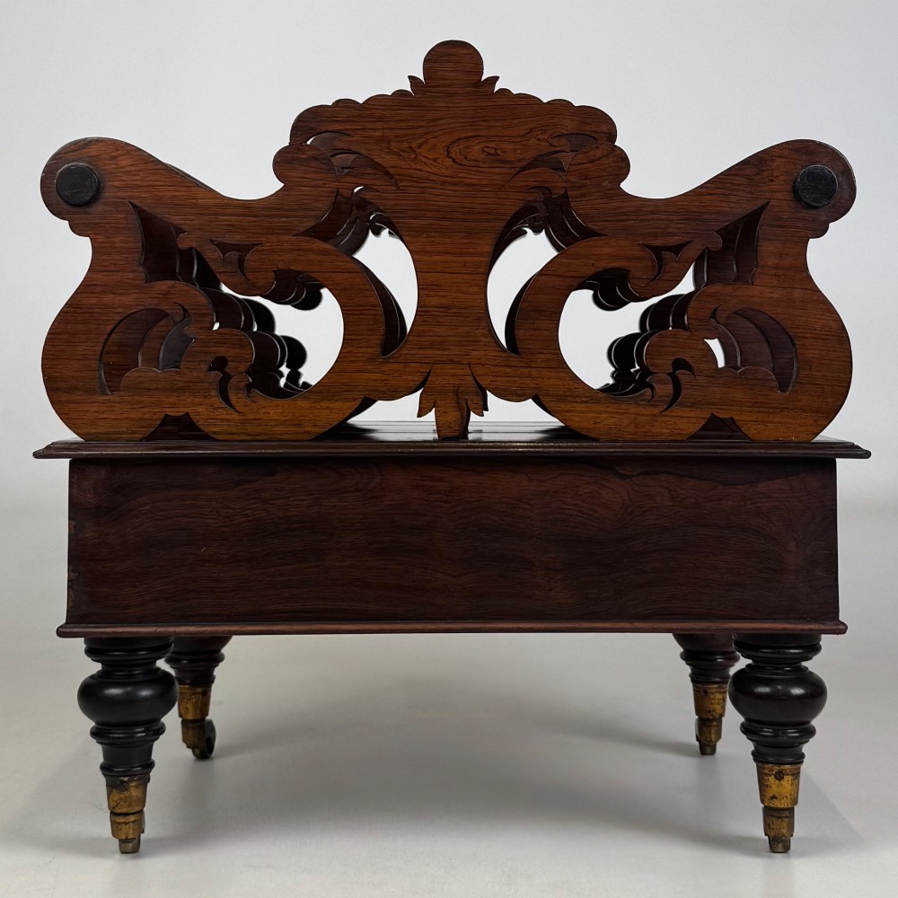 A dark rosewood Victorian canterbury with central acanthus carving, double scrollwork dividers, a single lower drawer, turned legs, and brass castors. View V