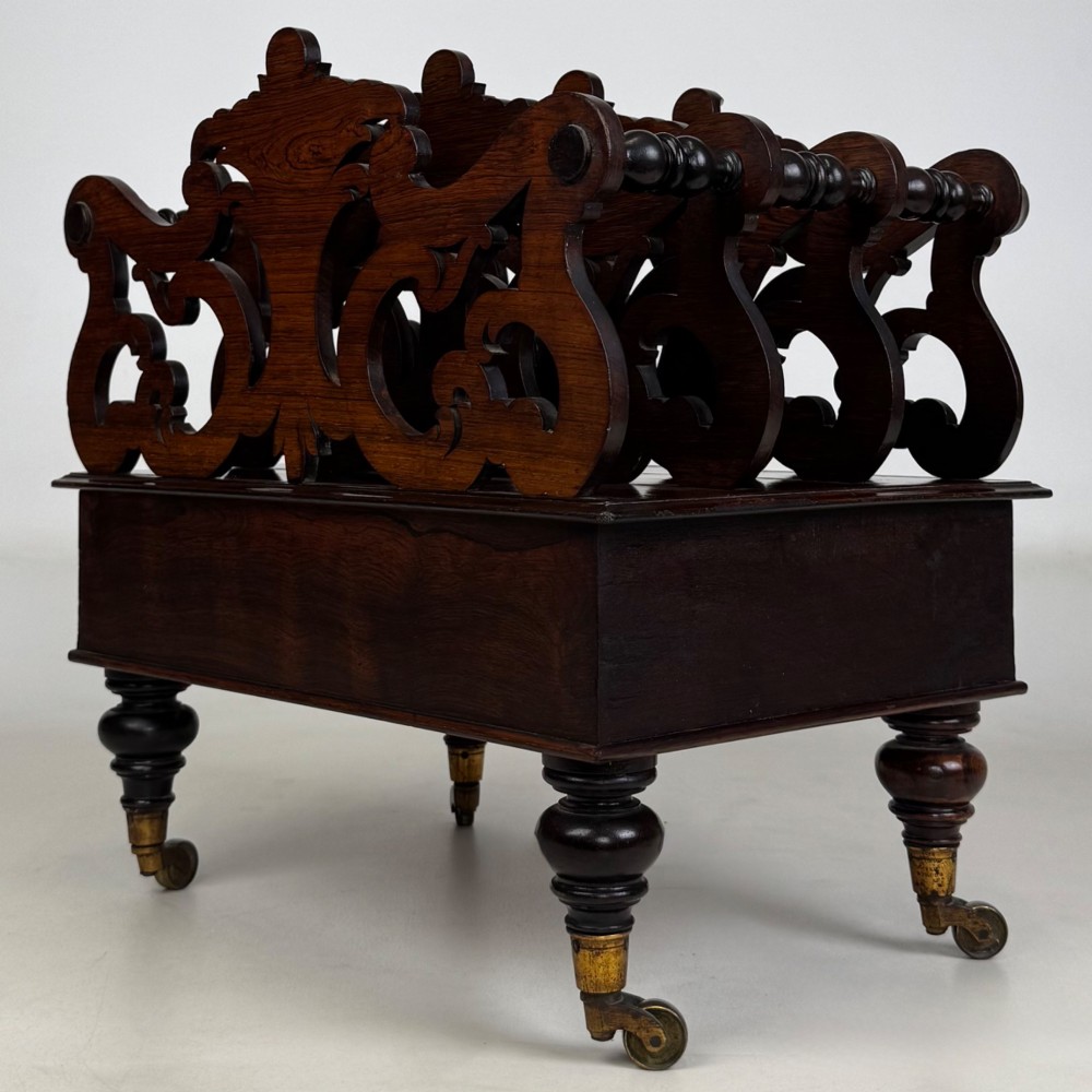 A dark rosewood Victorian canterbury with central acanthus carving, double scrollwork dividers, a single lower drawer, turned legs, and brass castors. View VI