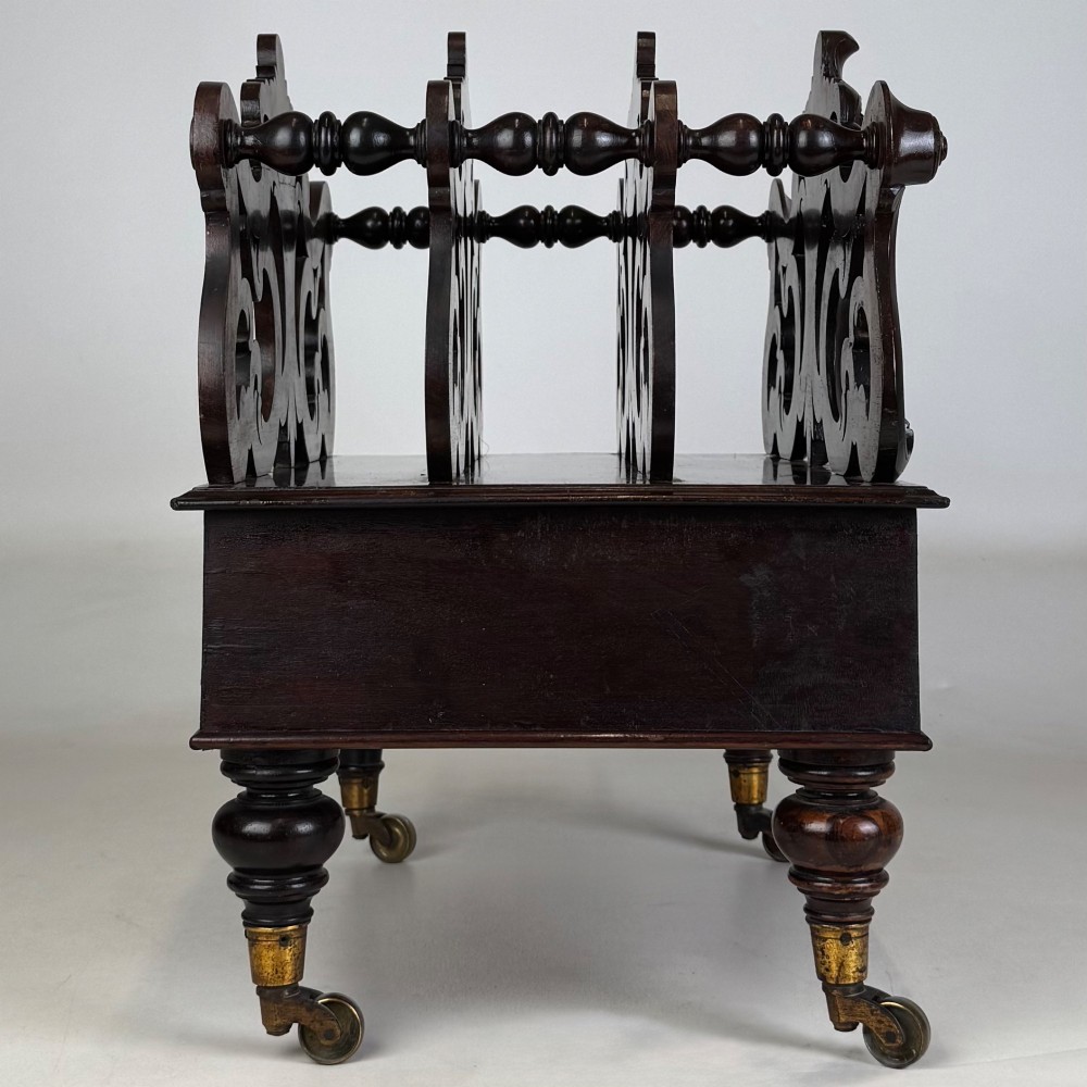A dark rosewood Victorian canterbury with central acanthus carving, double scrollwork dividers, a single lower drawer, turned legs, and brass castors. View VII