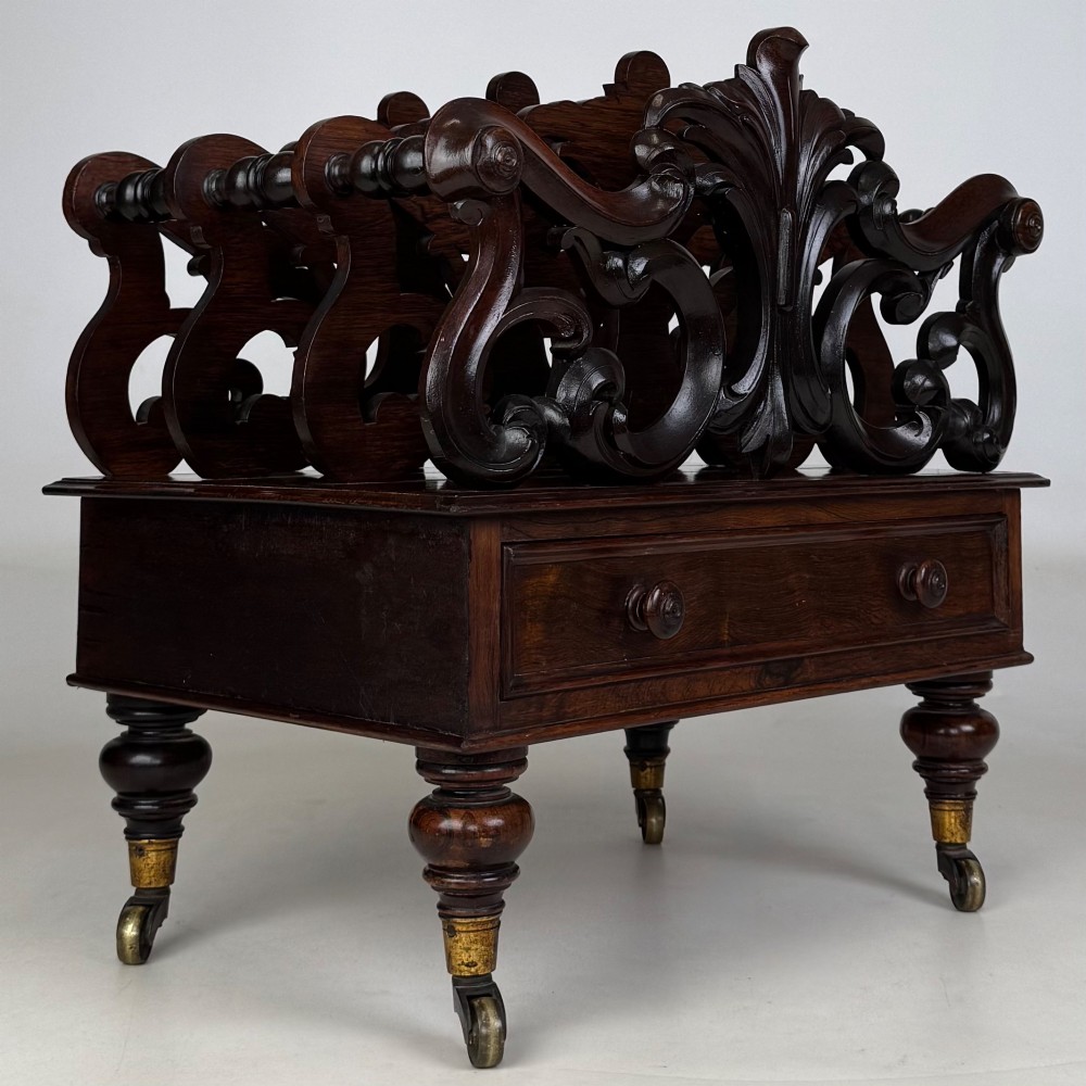 A dark rosewood Victorian canterbury with central acanthus carving, double scrollwork dividers, a single lower drawer, turned legs, and brass castors. View VIII