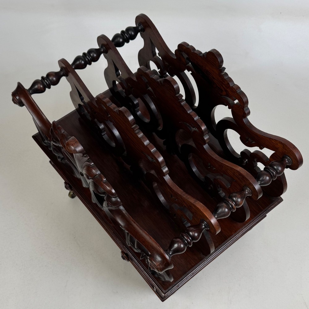 A dark rosewood Victorian canterbury with central acanthus carving, double scrollwork dividers, a single lower drawer, turned legs, and brass castors. View IX