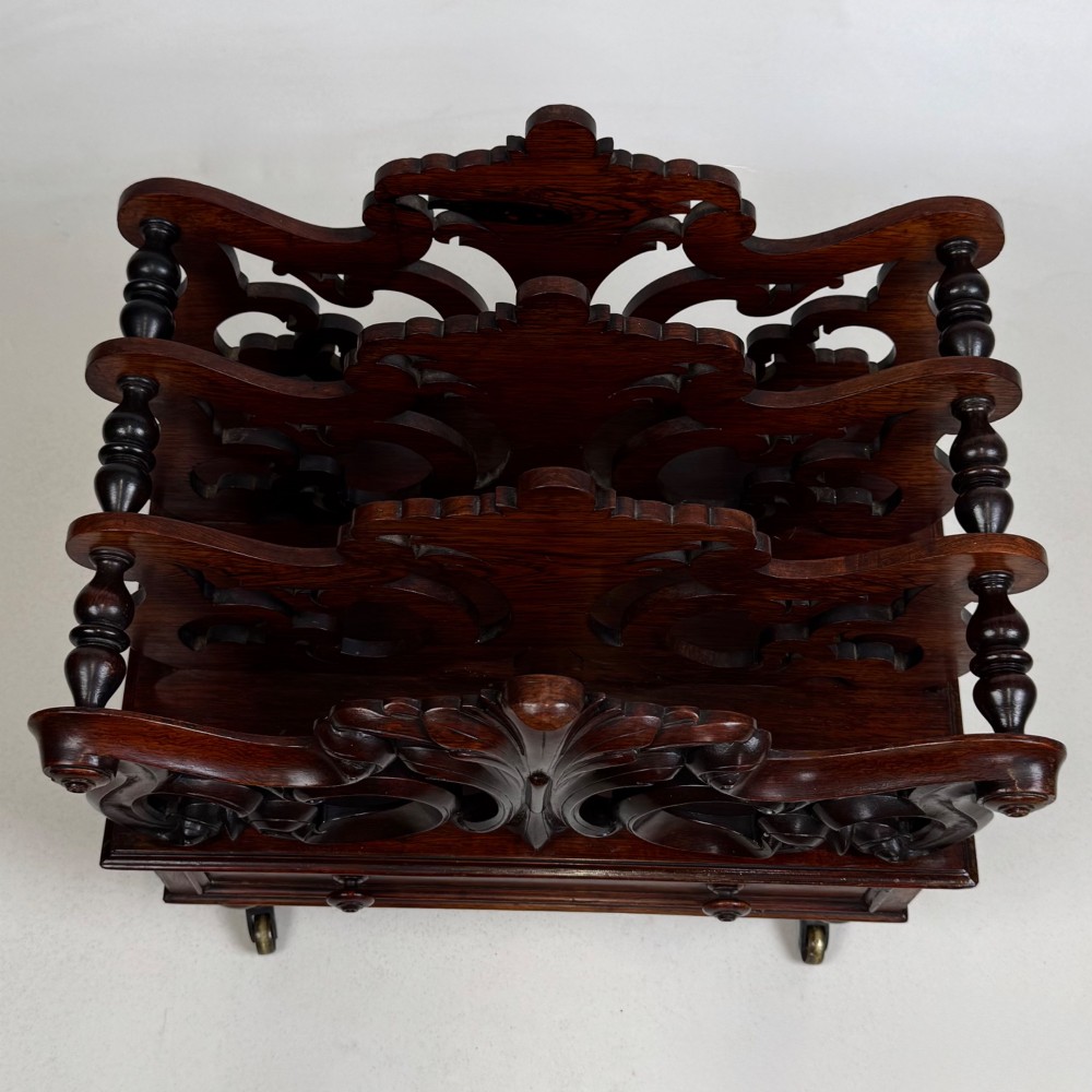 A dark rosewood Victorian canterbury with central acanthus carving, double scrollwork dividers, a single lower drawer, turned legs, and brass castors. View X