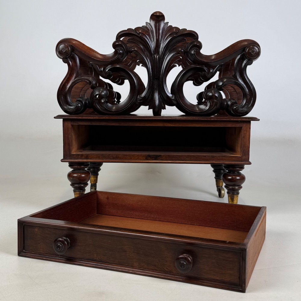 A dark rosewood Victorian canterbury with central acanthus carving, double scrollwork dividers, a single lower drawer, turned legs, and brass castors. View XI