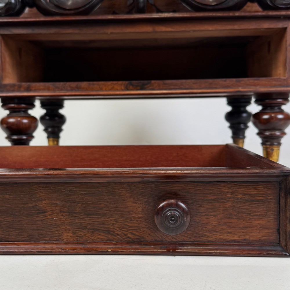 A dark rosewood Victorian canterbury with central acanthus carving, double scrollwork dividers, a single lower drawer, turned legs, and brass castors. View XII