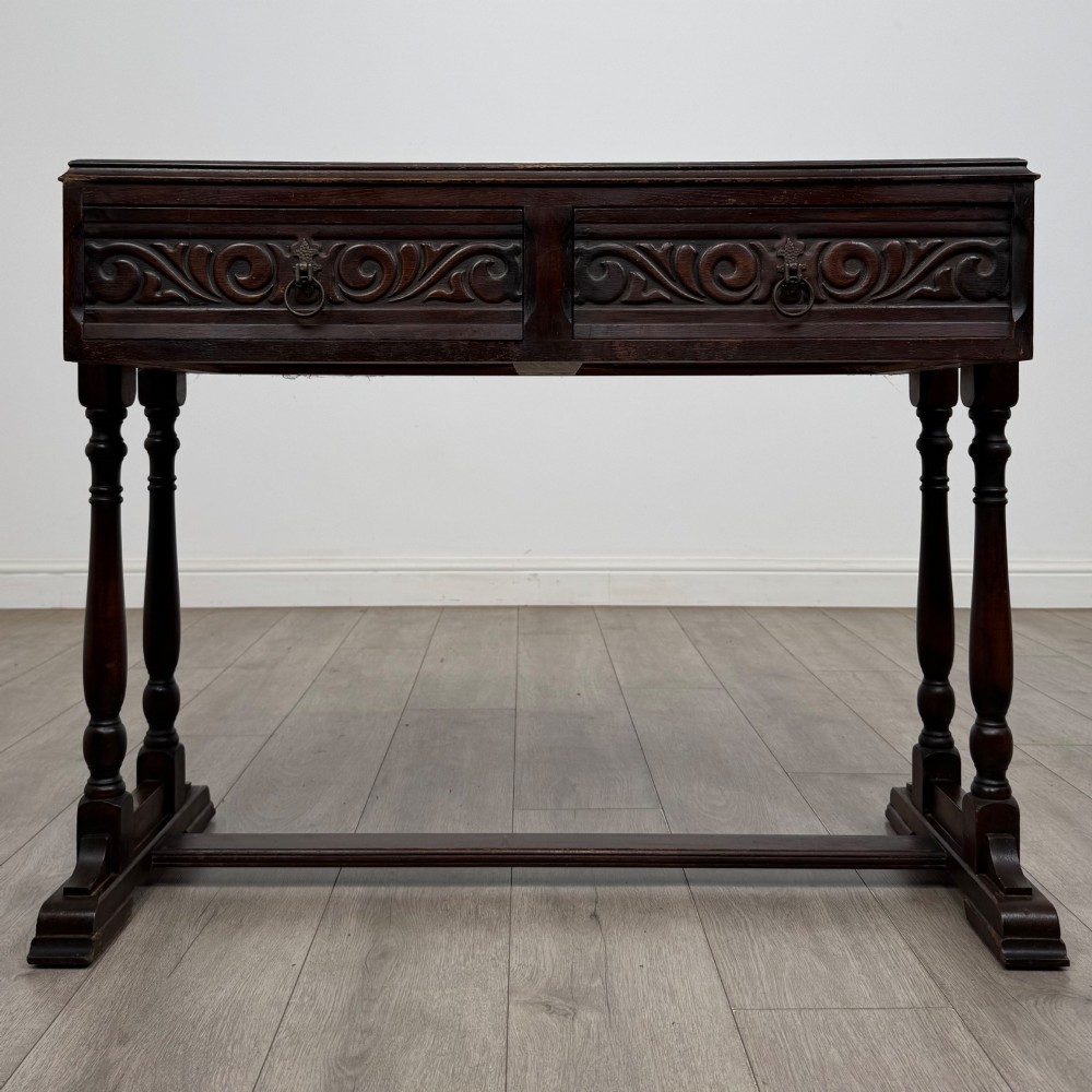 A dark brown carved oak side table featuring two scroll-carved front drawers with metal drop handles and turned trestle legs.