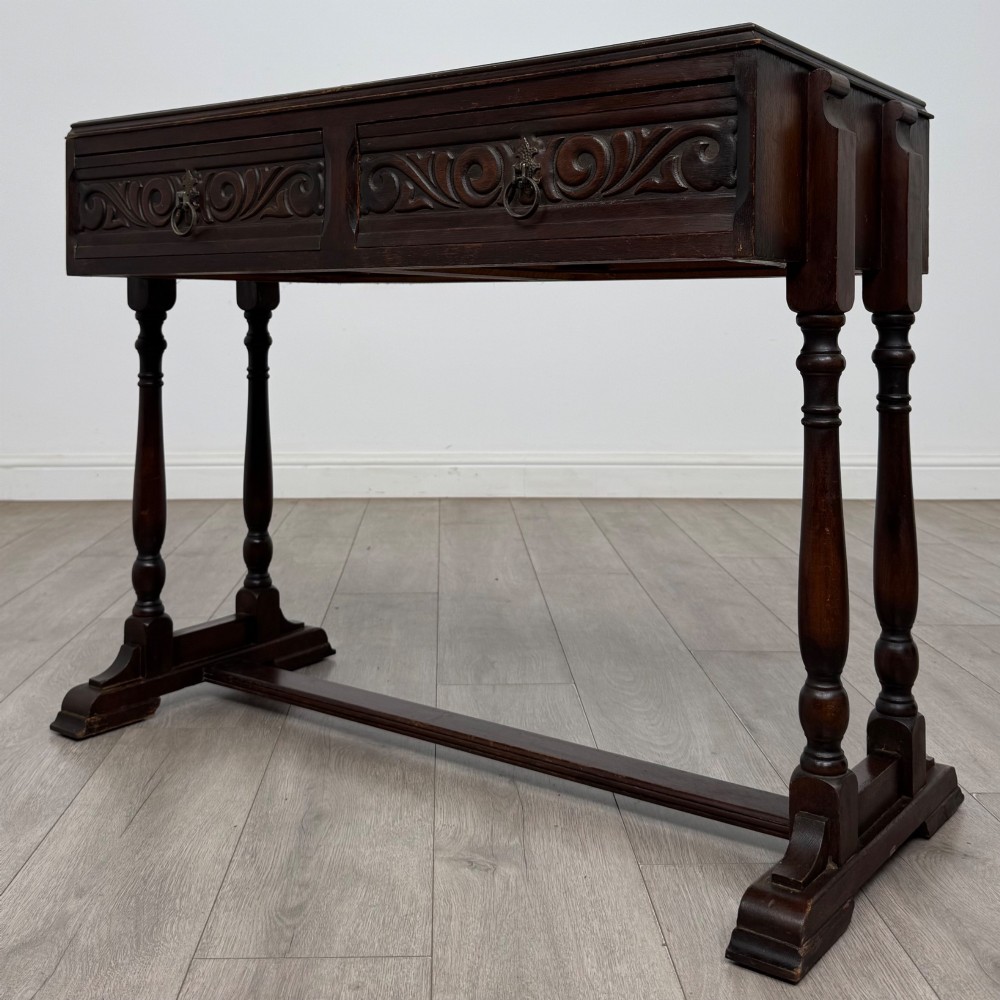 A dark brown carved oak side table featuring two scroll-carved front drawers with metal drop handles and turned trestle legs. View II