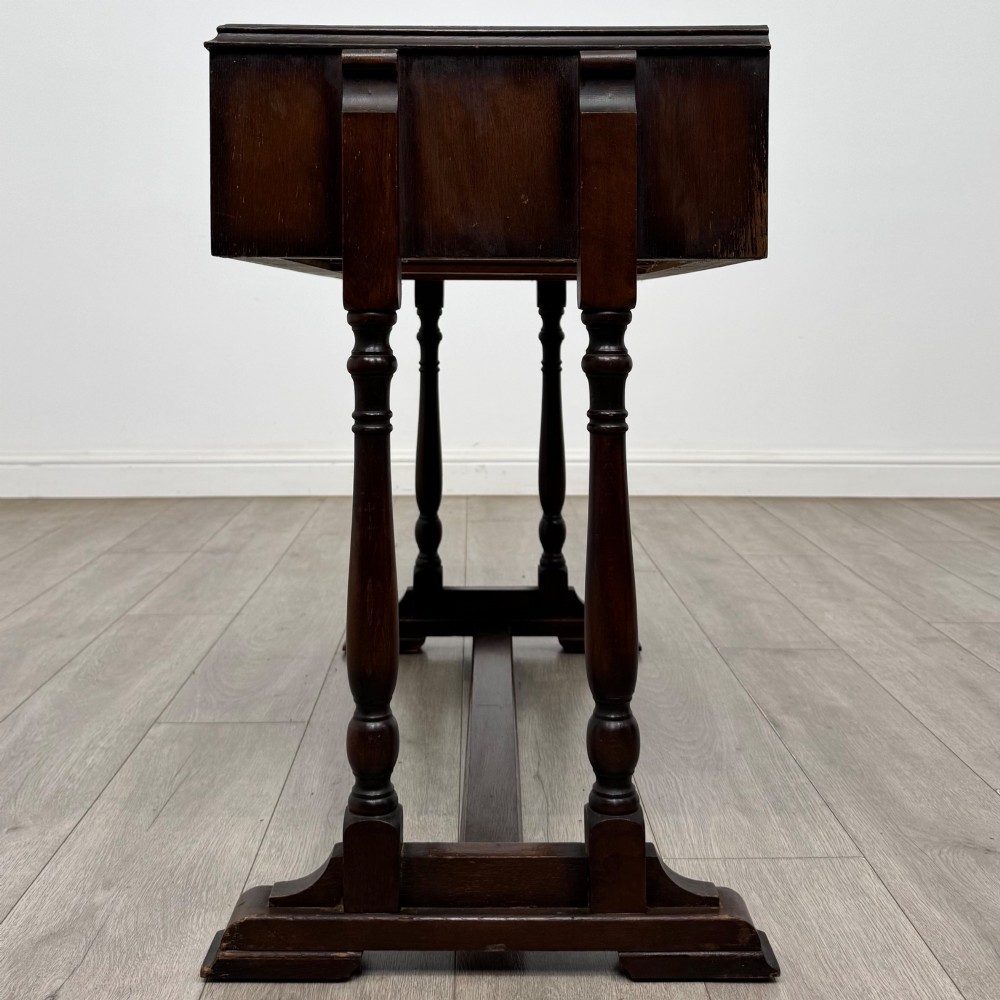 A dark brown carved oak side table featuring two scroll-carved front drawers with metal drop handles and turned trestle legs. View III