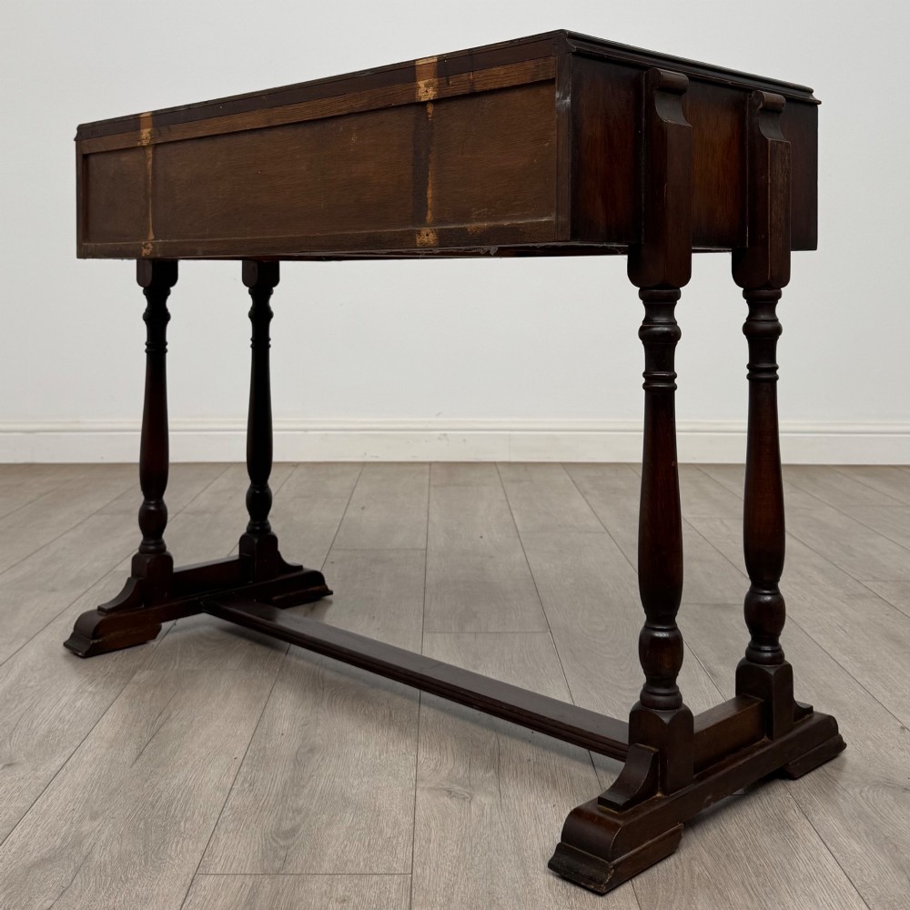 A dark brown carved oak side table featuring two scroll-carved front drawers with metal drop handles and turned trestle legs. View VI