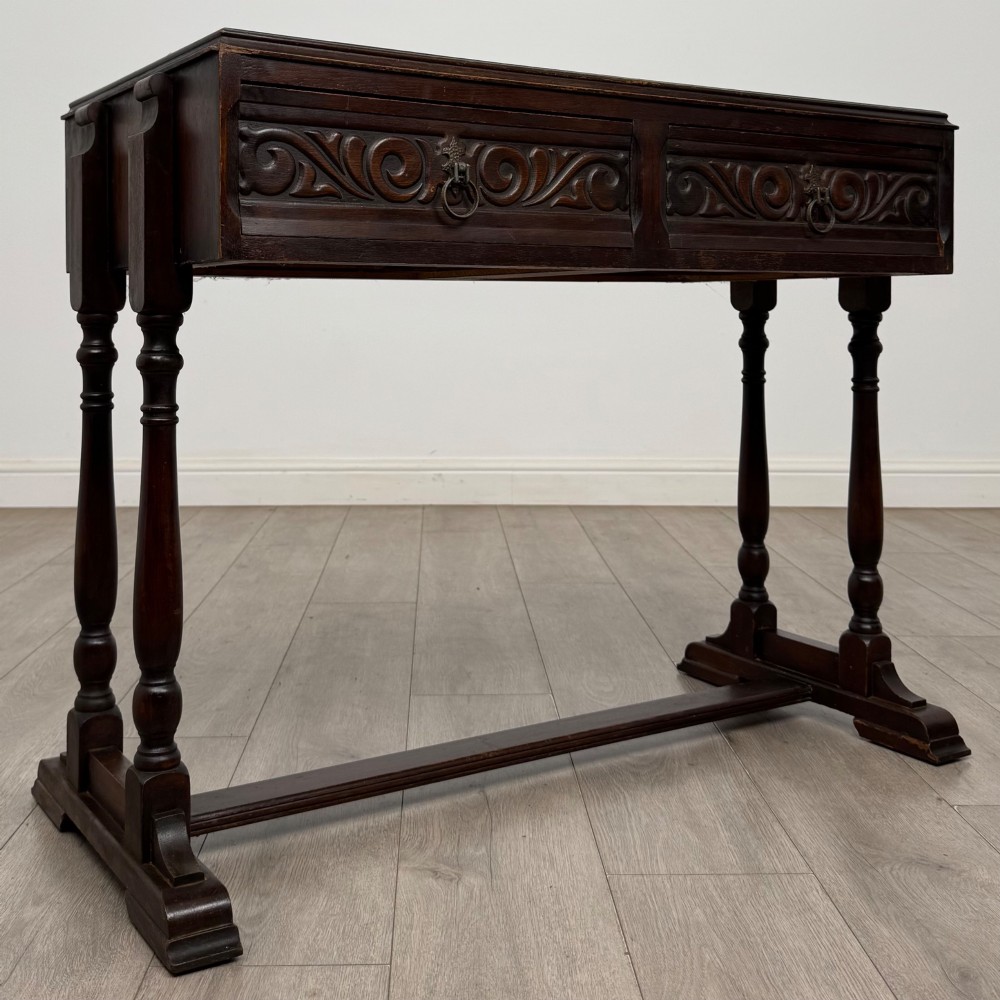 A dark brown carved oak side table featuring two scroll-carved front drawers with metal drop handles and turned trestle legs. View VIII