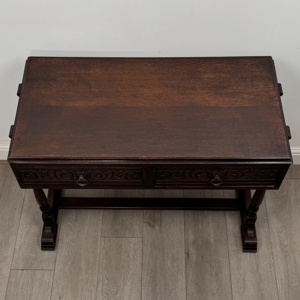 A dark brown carved oak side table featuring two scroll-carved front drawers with metal drop handles and turned trestle legs. View IX