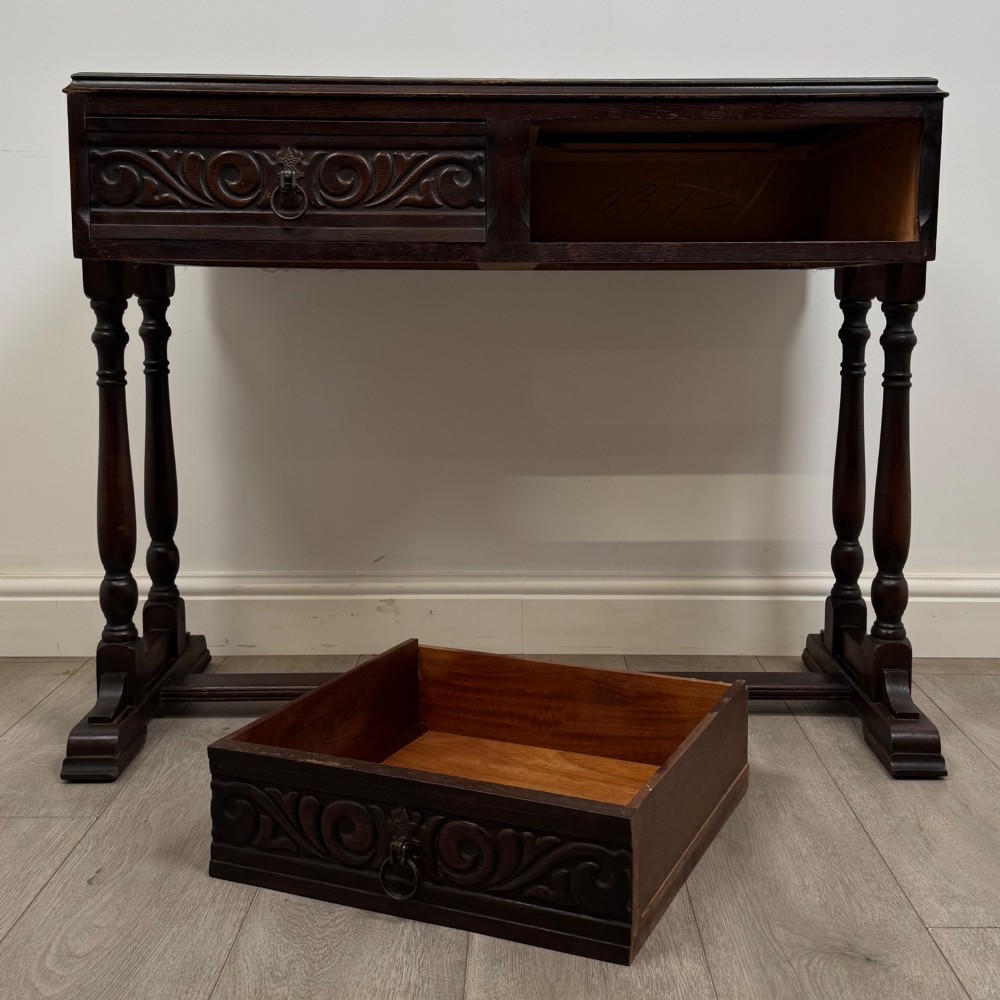 A dark brown carved oak side table featuring two scroll-carved front drawers with metal drop handles and turned trestle legs. View X
