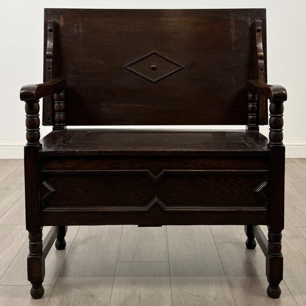 A dark oak monks bench featuring a panelled backrest with a central diamond motif, turned arm supports, and a lift-up seat.