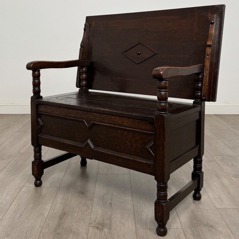 A dark oak monks bench featuring a panelled backrest with a central diamond motif, turned arm supports, and a lift-up seat. View III