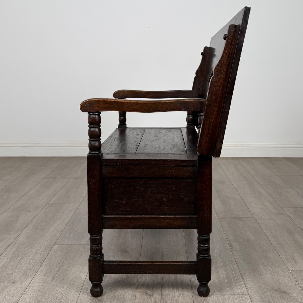 A dark oak monks bench featuring a panelled backrest with a central diamond motif, turned arm supports, and a lift-up seat. View IV