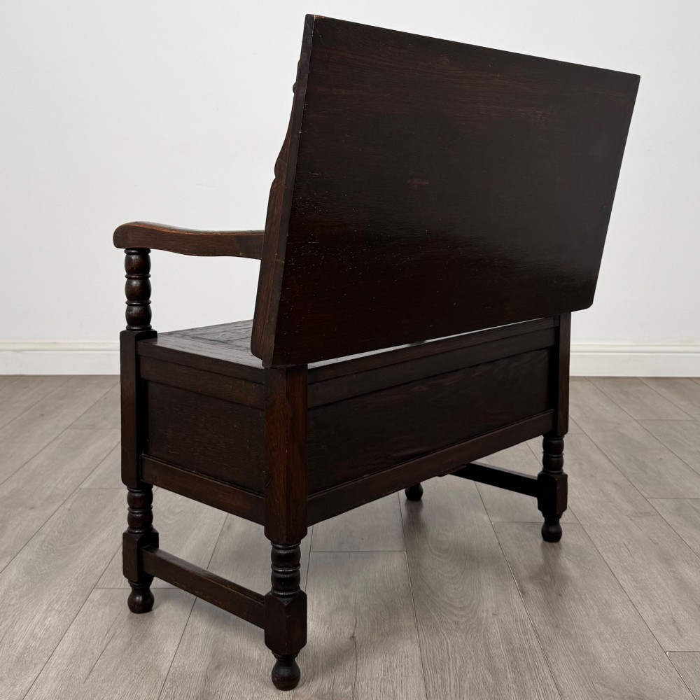 A dark oak monks bench featuring a panelled backrest with a central diamond motif, turned arm supports, and a lift-up seat. View V