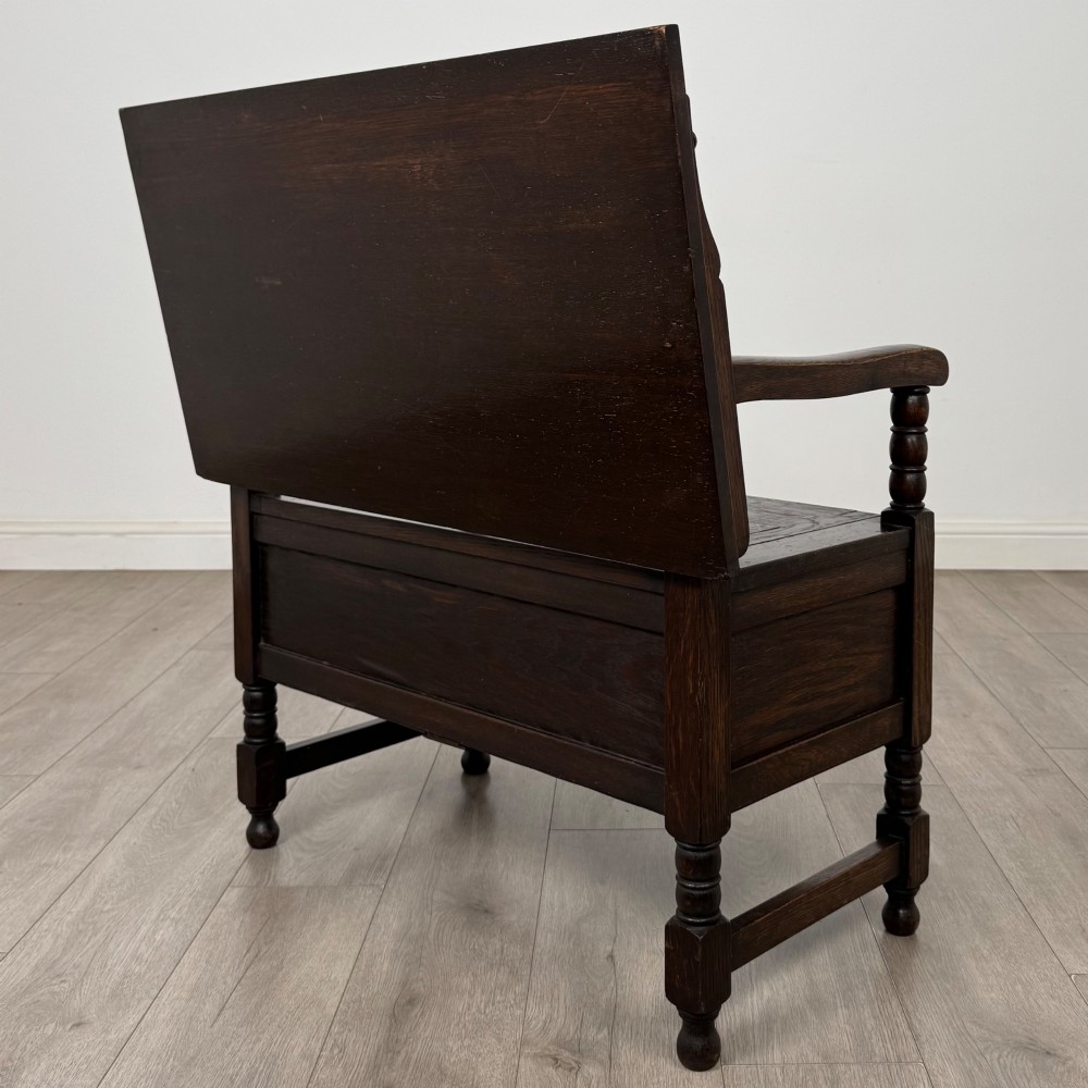 A dark oak monks bench featuring a panelled backrest with a central diamond motif, turned arm supports, and a lift-up seat. View VII