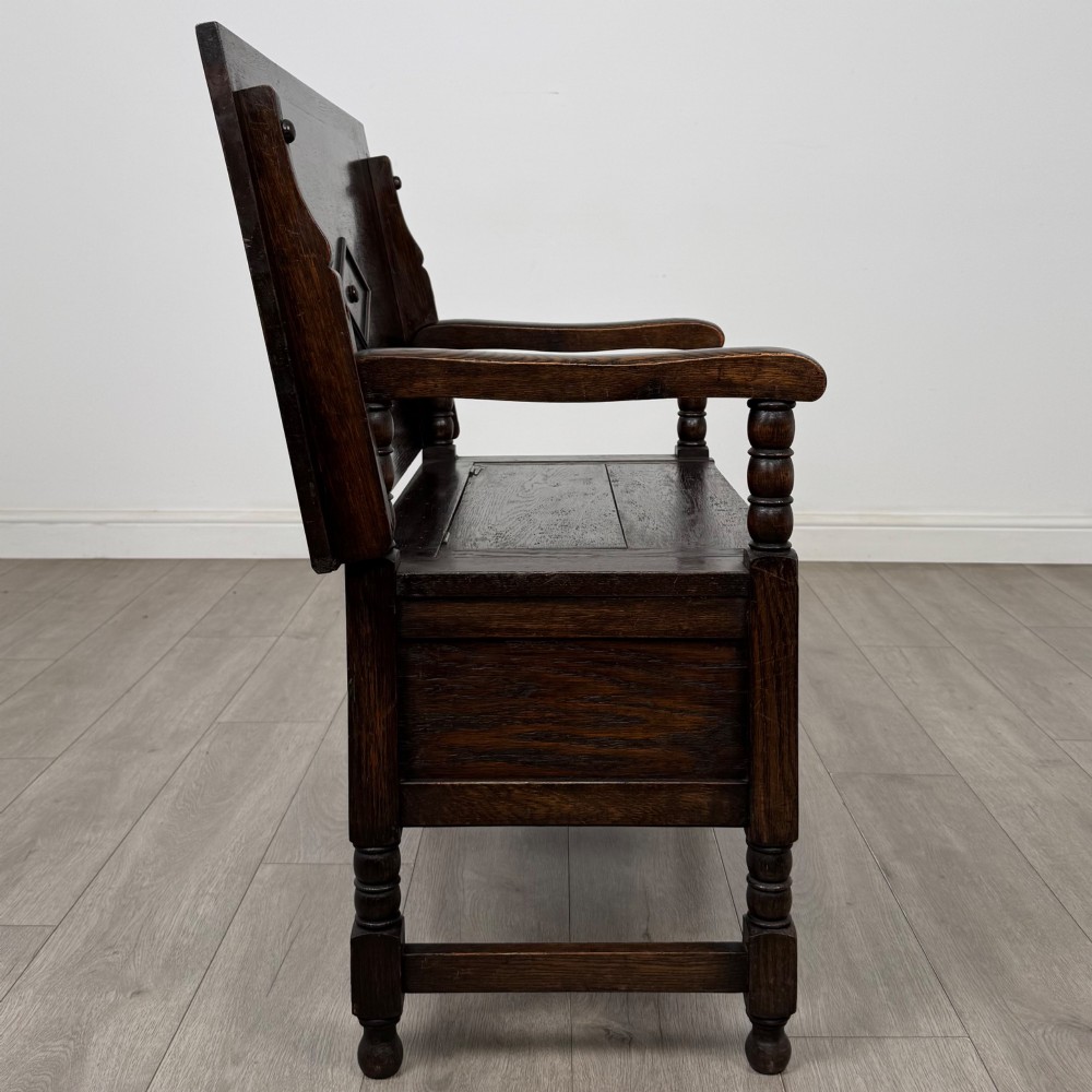 A dark oak monks bench featuring a panelled backrest with a central diamond motif, turned arm supports, and a lift-up seat. View VIII
