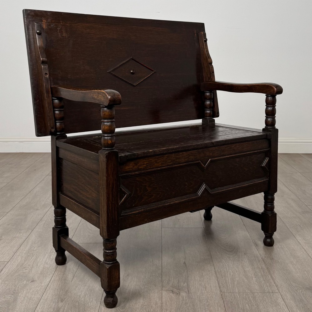 A dark oak monks bench featuring a panelled backrest with a central diamond motif, turned arm supports, and a lift-up seat. View IX