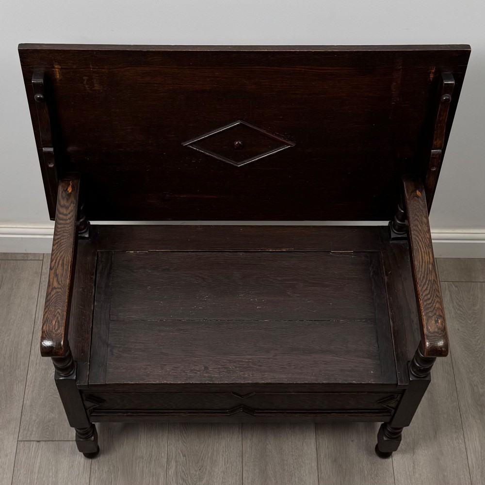 A dark oak monks bench featuring a panelled backrest with a central diamond motif, turned arm supports, and a lift-up seat. View X