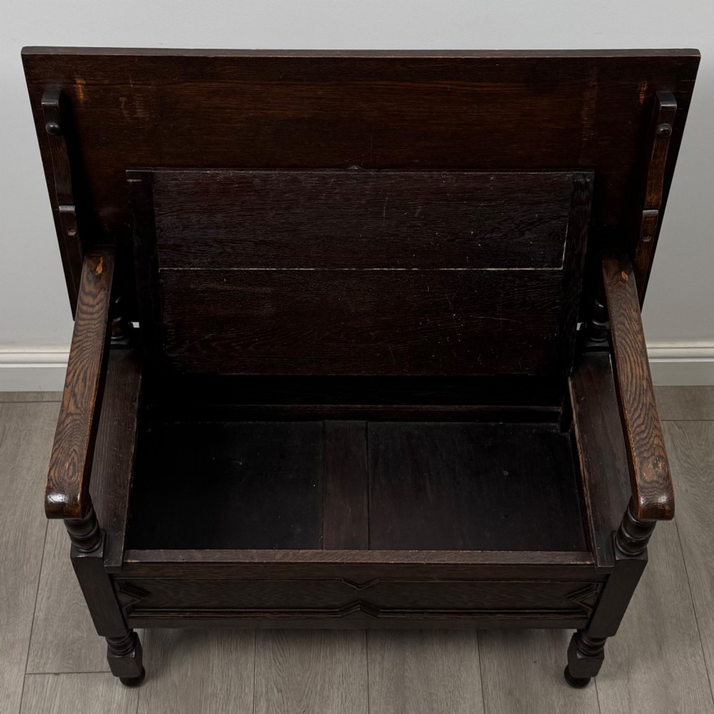 A dark oak monks bench featuring a panelled backrest with a central diamond motif, turned arm supports, and a lift-up seat. View XI