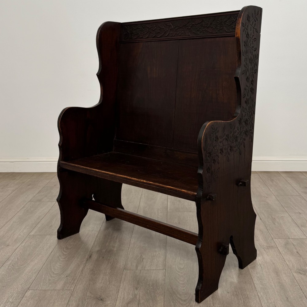 A dark oak high-back settle featuring a floral relief carving along the top rail, shaped side panels, and a stretcher base. View II
