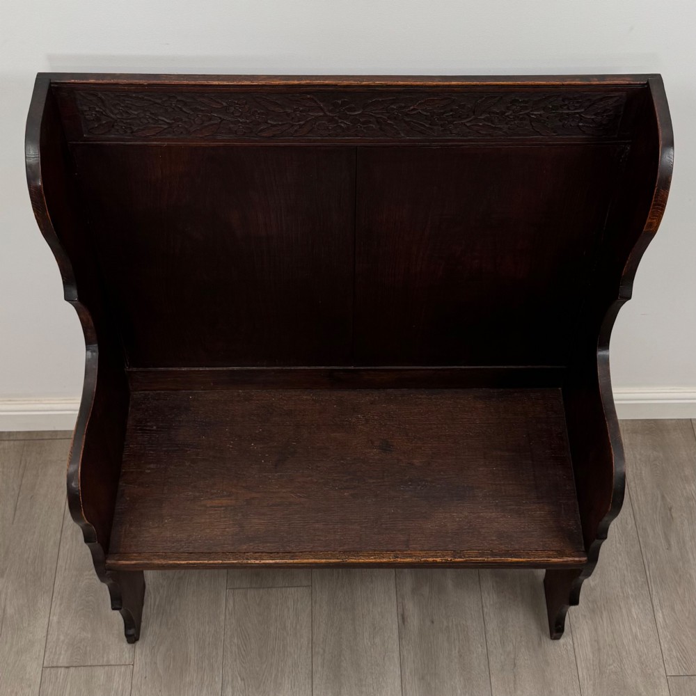 A dark oak high-back settle featuring a floral relief carving along the top rail, shaped side panels, and a stretcher base. View IX
