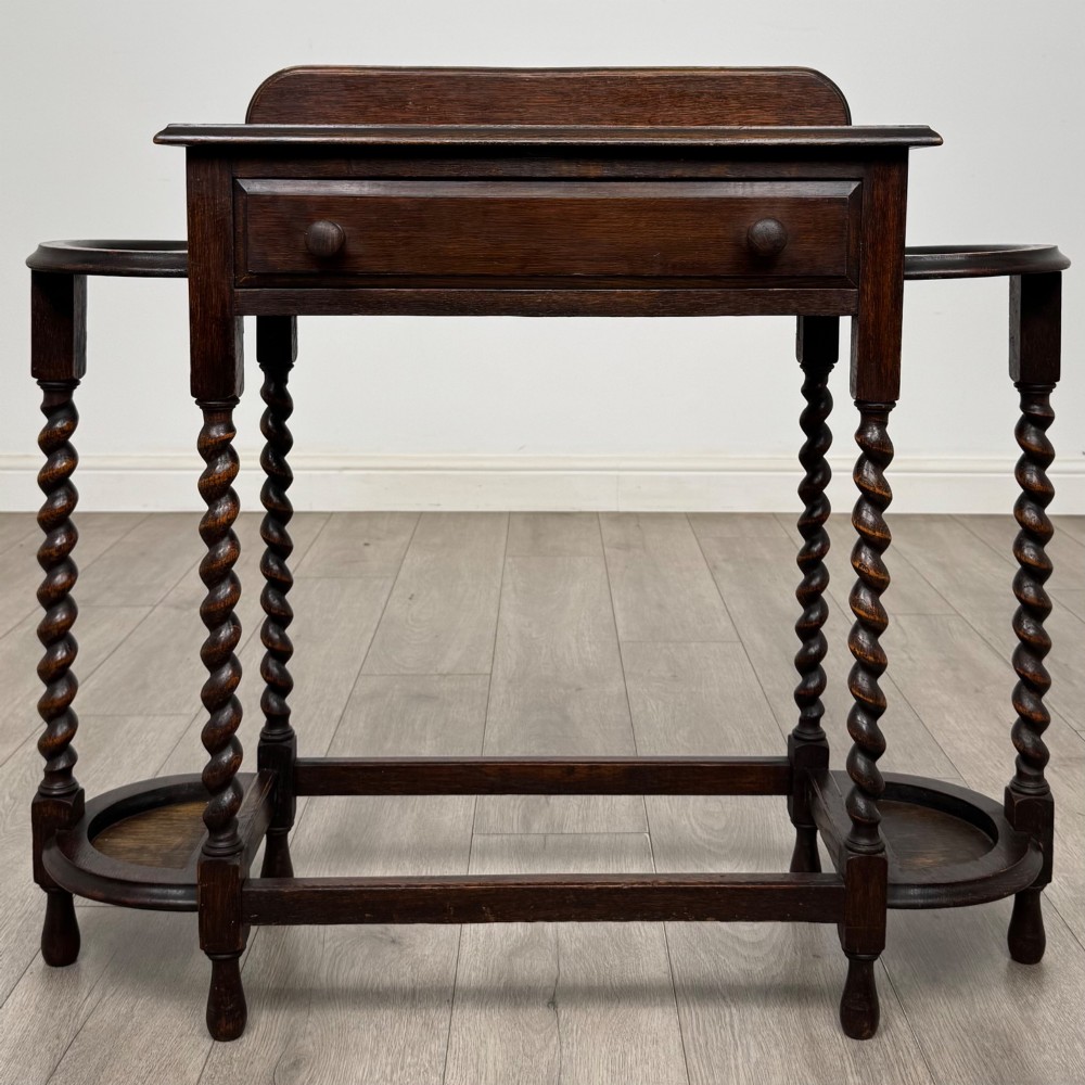 A dark oak Edwardian hall stand featuring turned barley twist legs, a central drawer with wooden knobs, and built-in side umbrella holders with drip trays.