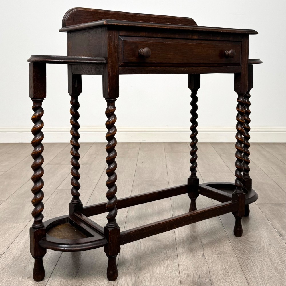 A dark oak Edwardian hall stand featuring turned barley twist legs, a central drawer with wooden knobs, and built-in side umbrella holders with drip trays. View VIII