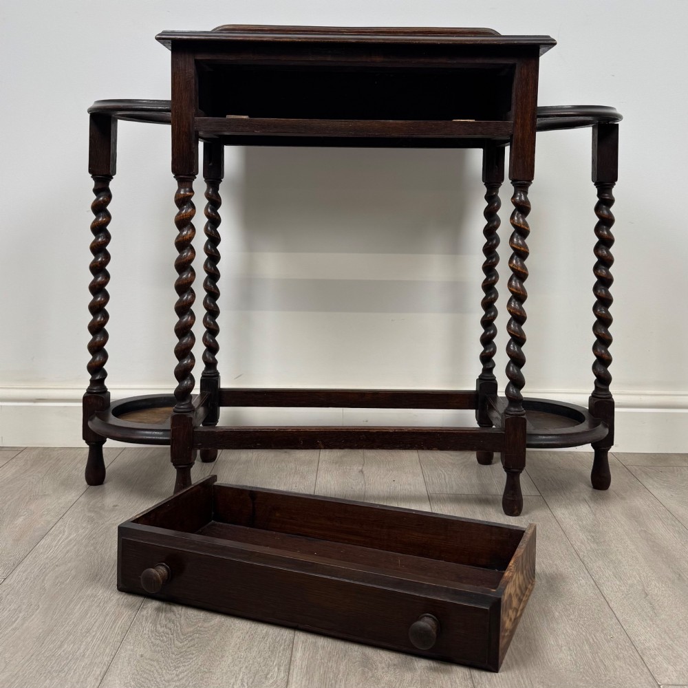A dark oak Edwardian hall stand featuring turned barley twist legs, a central drawer with wooden knobs, and built-in side umbrella holders with drip trays. View X