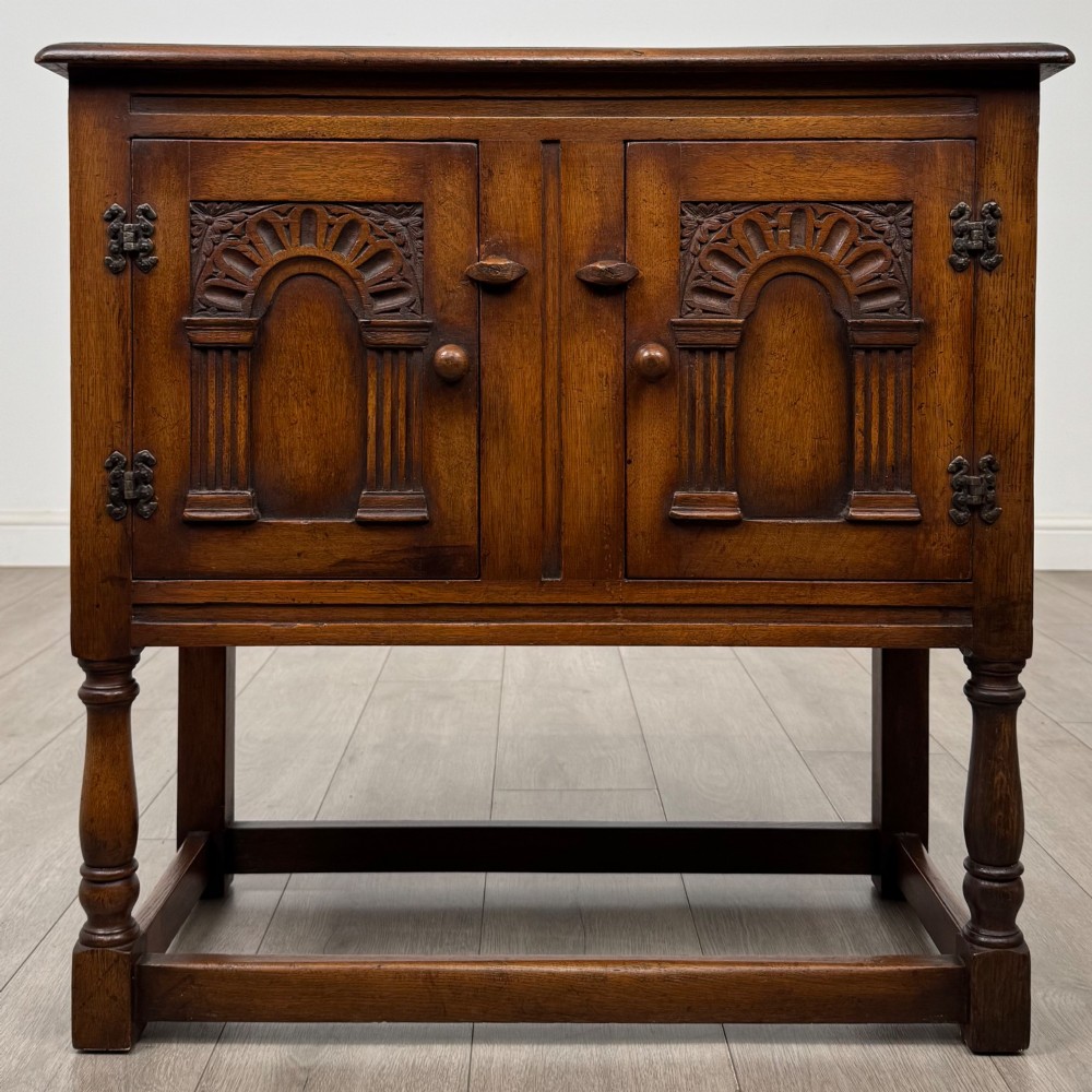 A small warm brown carved oak cupboard with double doors featuring carved arches, iron hinges, turned legs, and a lower stretcher base.