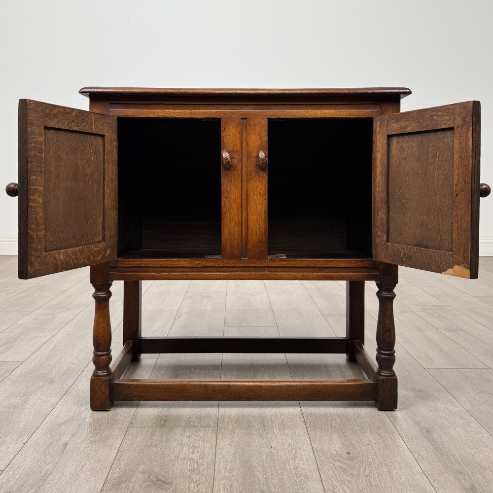 A small warm brown carved oak cupboard with double doors featuring carved arches, iron hinges, turned legs, and a lower stretcher base. View II