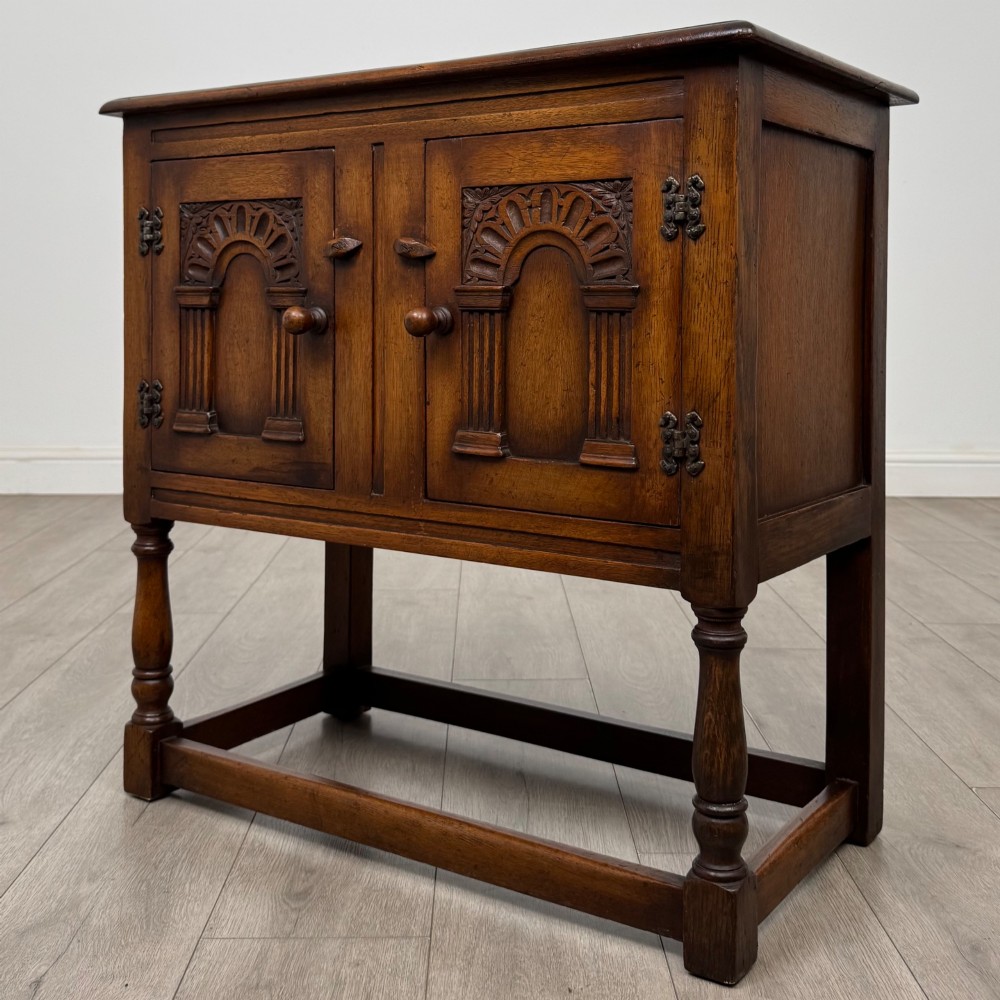 A small warm brown carved oak cupboard with double doors featuring carved arches, iron hinges, turned legs, and a lower stretcher base. View III