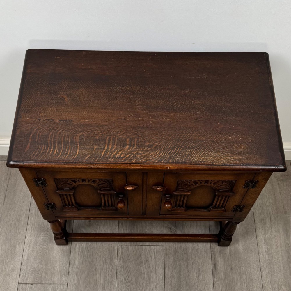 A small warm brown carved oak cupboard with double doors featuring carved arches, iron hinges, turned legs, and a lower stretcher base. View X