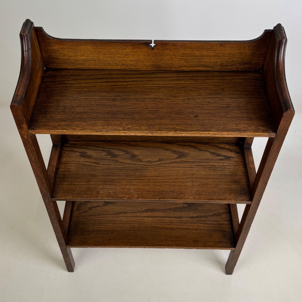 An early 20th-century Arts and Crafts solid oak open bookcase featuring three fixed shelves and a shaped gallery backboard with a cutout heart motif. View X