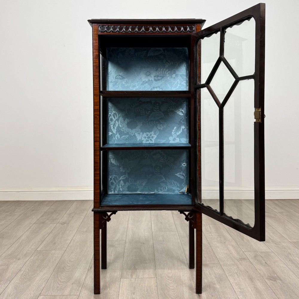 A slender Edwardian mahogany glazed display cabinet featuring astragal door bars and a fabric-lined interior raised on square legs. View II