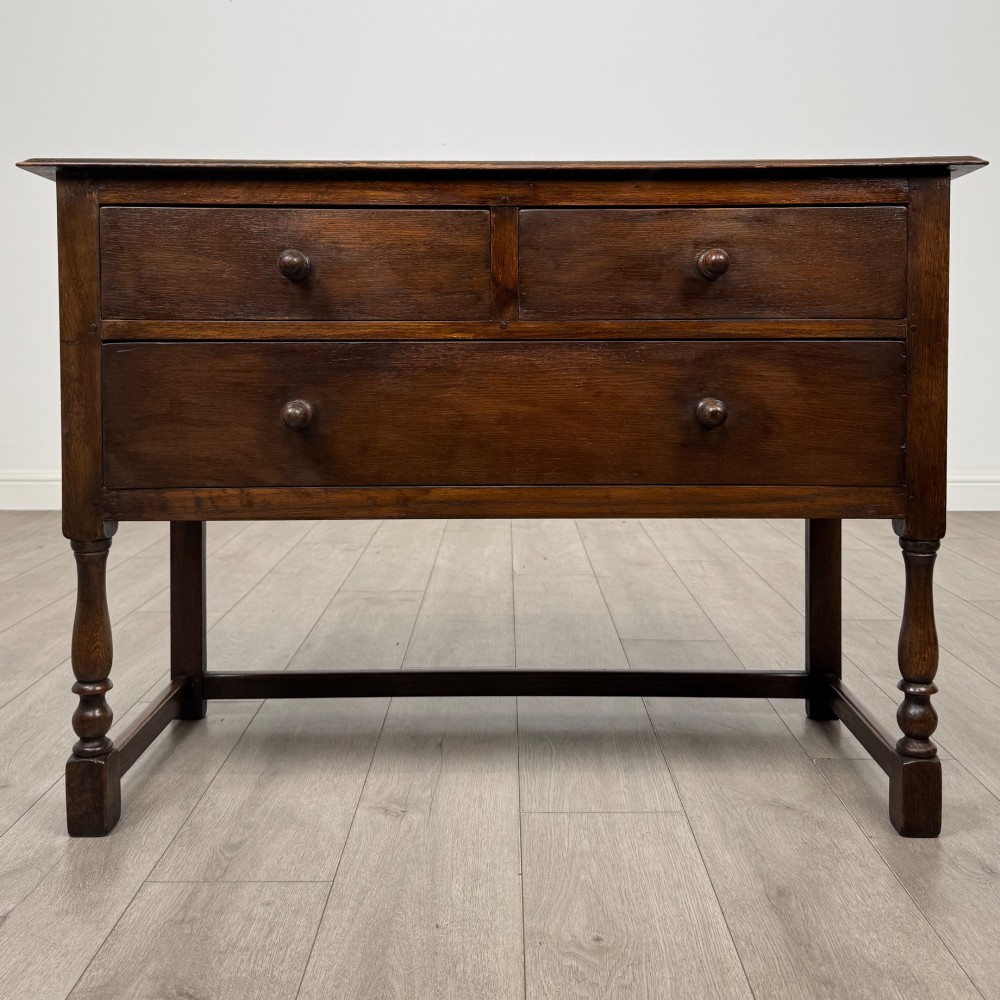A dark oak chest featuring two top drawers above one wide bottom drawer, resting on turned legs connected by a low stretcher frame.