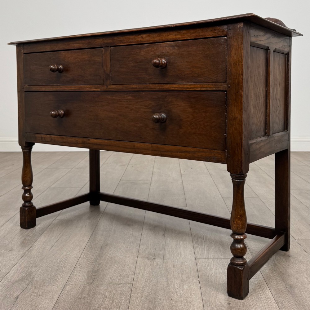 A dark oak chest featuring two top drawers above one wide bottom drawer, resting on turned legs connected by a low stretcher frame. View II