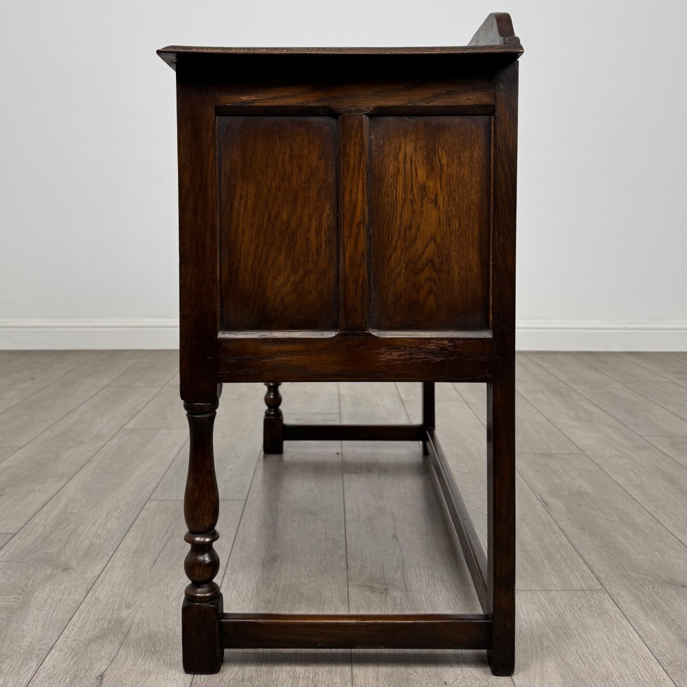 A dark oak chest featuring two top drawers above one wide bottom drawer, resting on turned legs connected by a low stretcher frame. View III