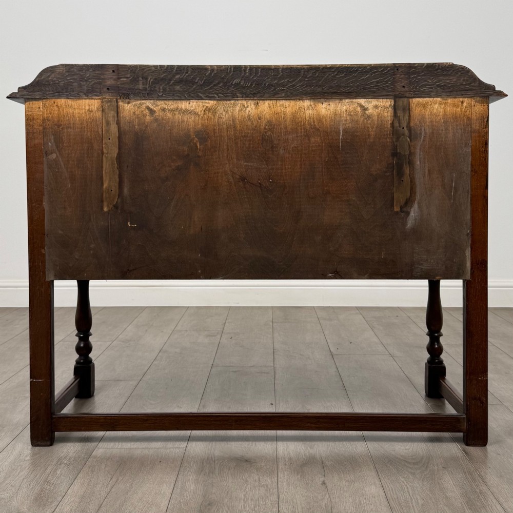 A dark oak chest featuring two top drawers above one wide bottom drawer, resting on turned legs connected by a low stretcher frame. View V