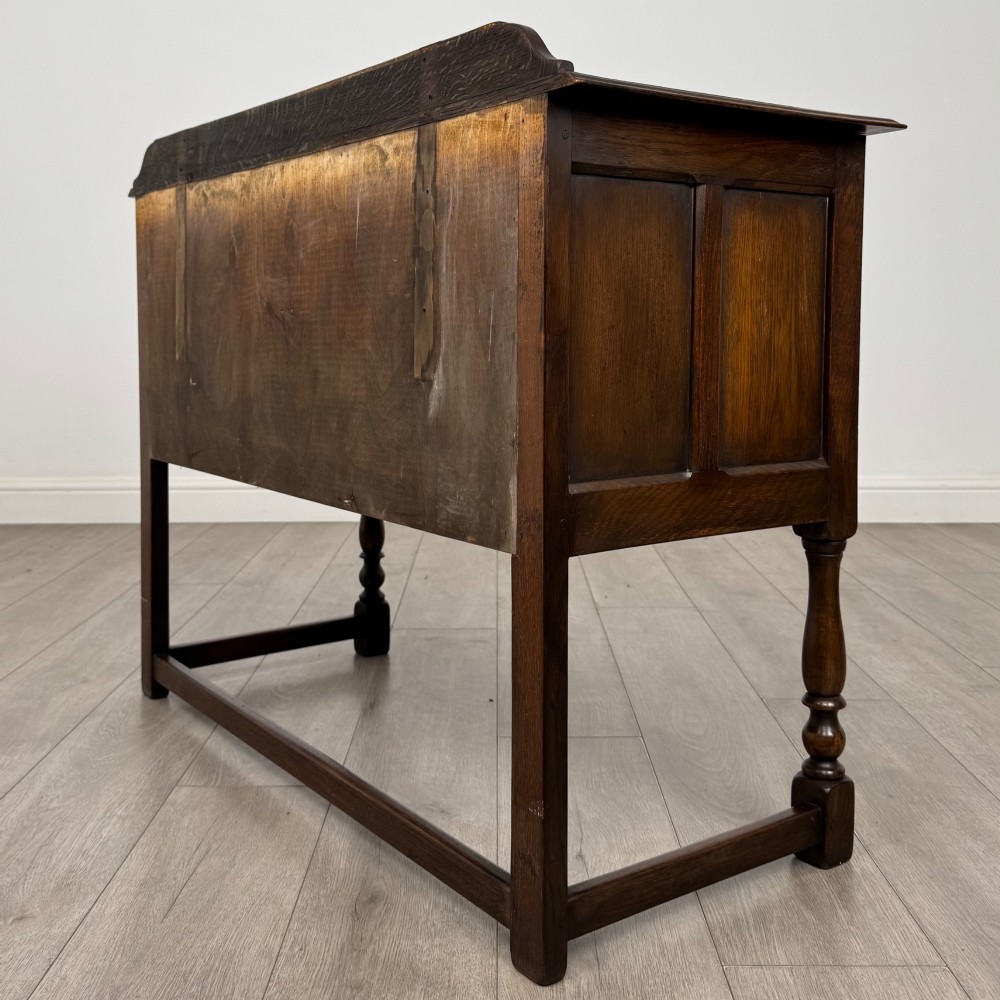 A dark oak chest featuring two top drawers above one wide bottom drawer, resting on turned legs connected by a low stretcher frame. View VI