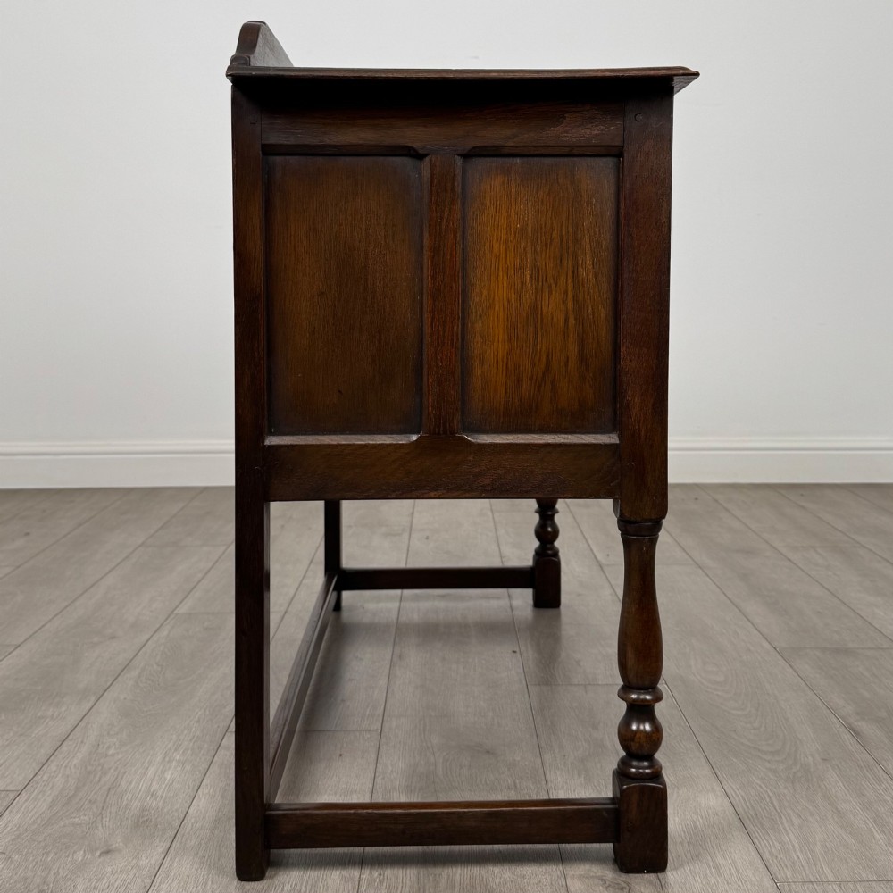 A dark oak chest featuring two top drawers above one wide bottom drawer, resting on turned legs connected by a low stretcher frame. View VII