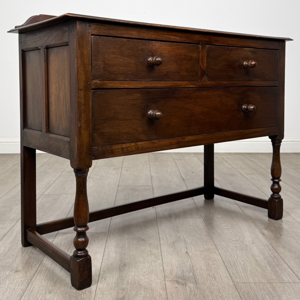 A dark oak chest featuring two top drawers above one wide bottom drawer, resting on turned legs connected by a low stretcher frame. View VIII