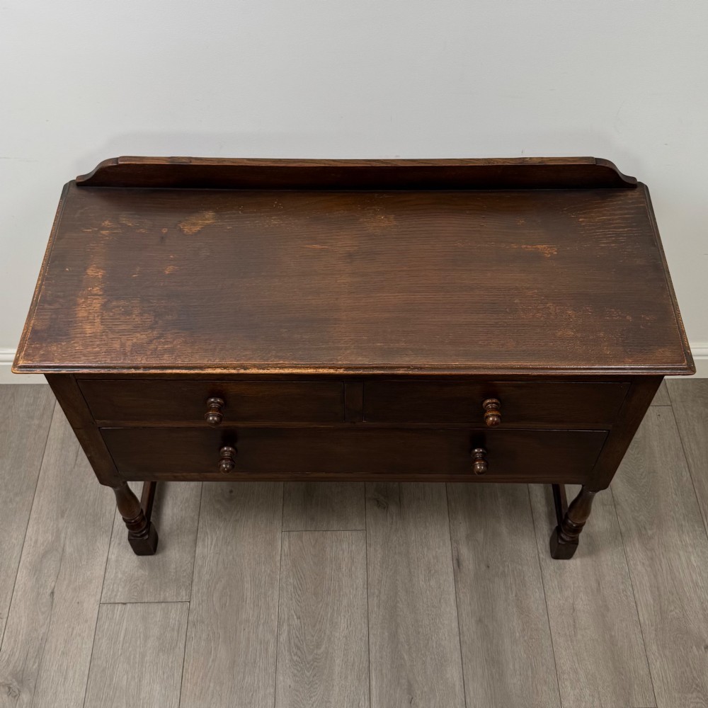 A dark oak chest featuring two top drawers above one wide bottom drawer, resting on turned legs connected by a low stretcher frame. View IX